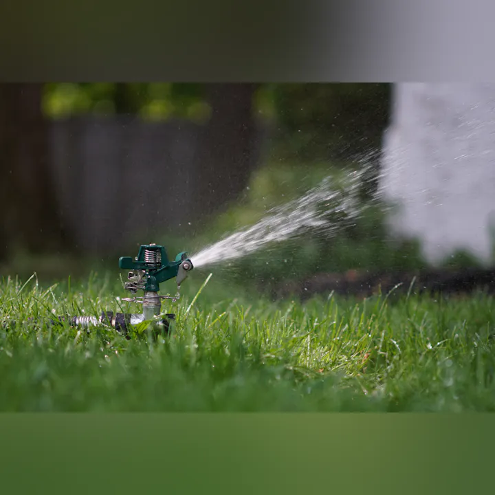 A green lawn sprinkler is spraying water over lush grass, creating droplets in the sunlight.
