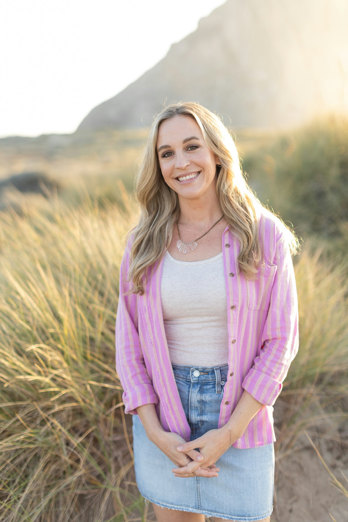 Smiling woman in a pink shirt and denim skirt standing in a grassy dune.