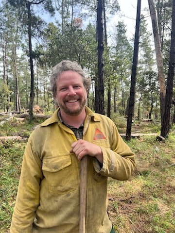 A smiling person in a forest wearing a yellow jacket, holding a tool, surrounded by tall trees.