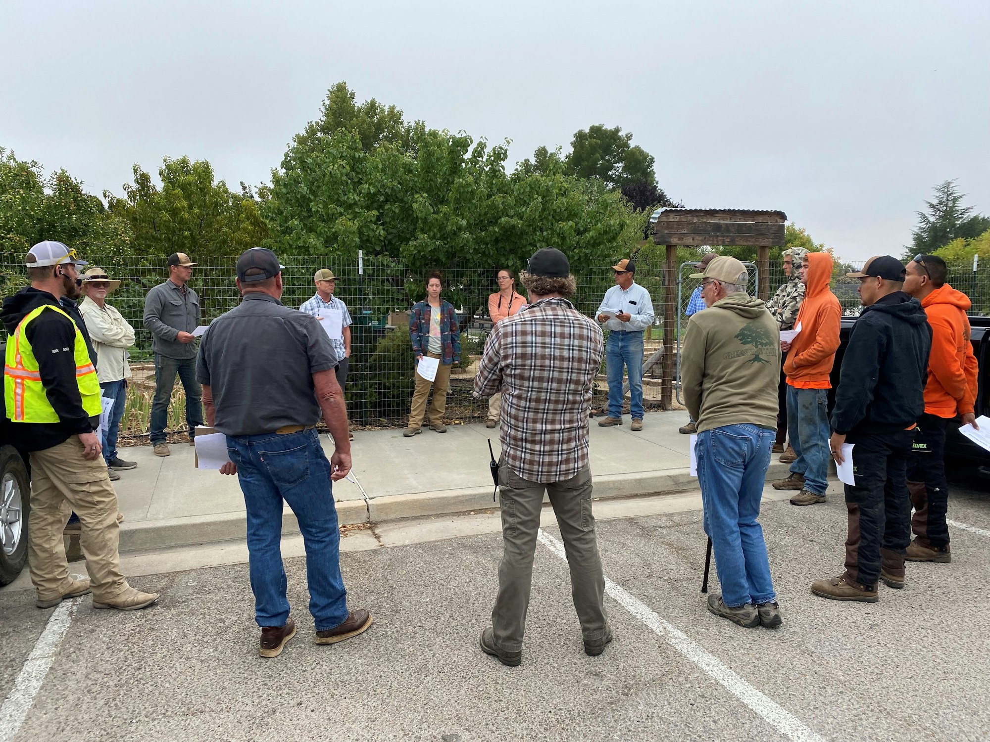 A group of people standing in a circle outdoors, some holding papers, possibly in a meeting or discussion.
