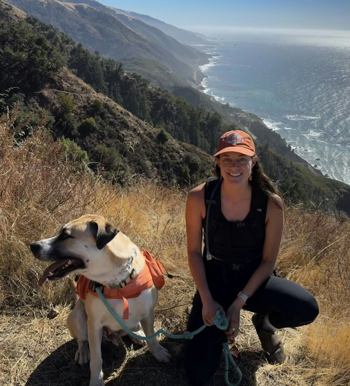 A woman kneels beside a large dog on a coastal hiking trail with the ocean and mountains in the background.