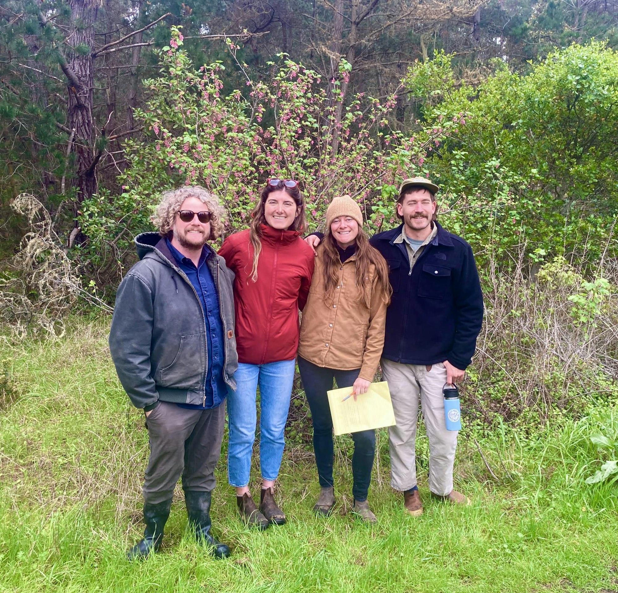 Four people posing outdoors with trees and shrubs in the background.