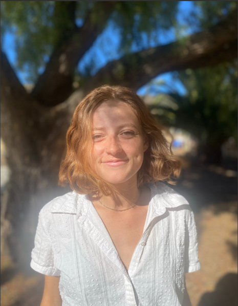A person with short hair smiles beneath a tree, wearing a white shirt, bathed in sunlight.