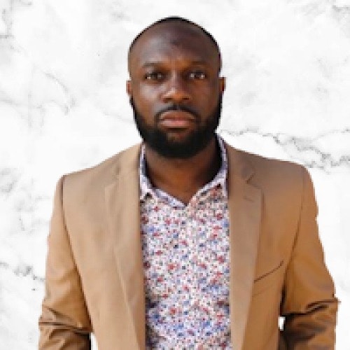 A man in a beige blazer and patterned shirt against a white marble background.