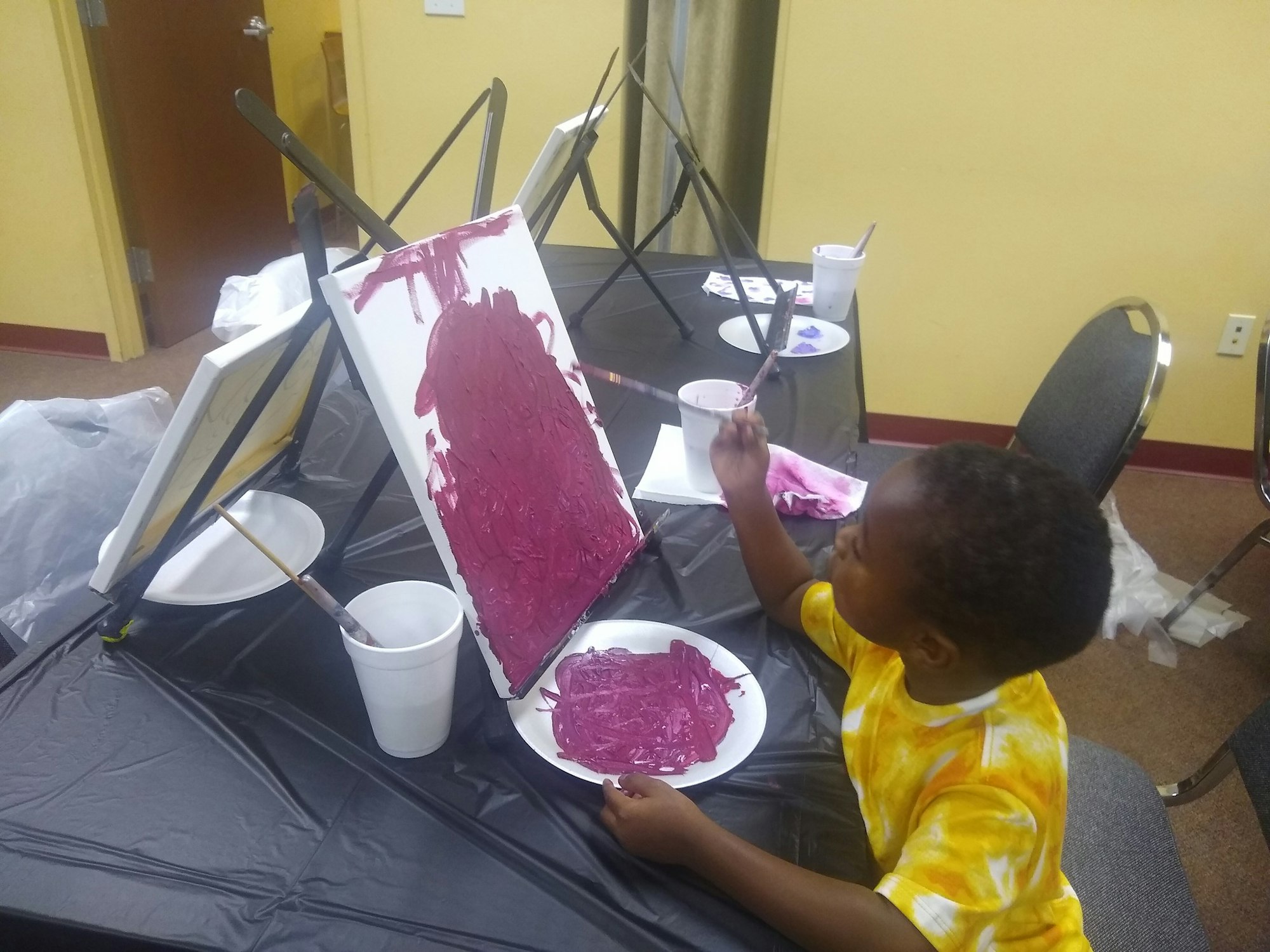 A child in a yellow shirt paints on a canvas, using a plate of purple paint, in an art room.