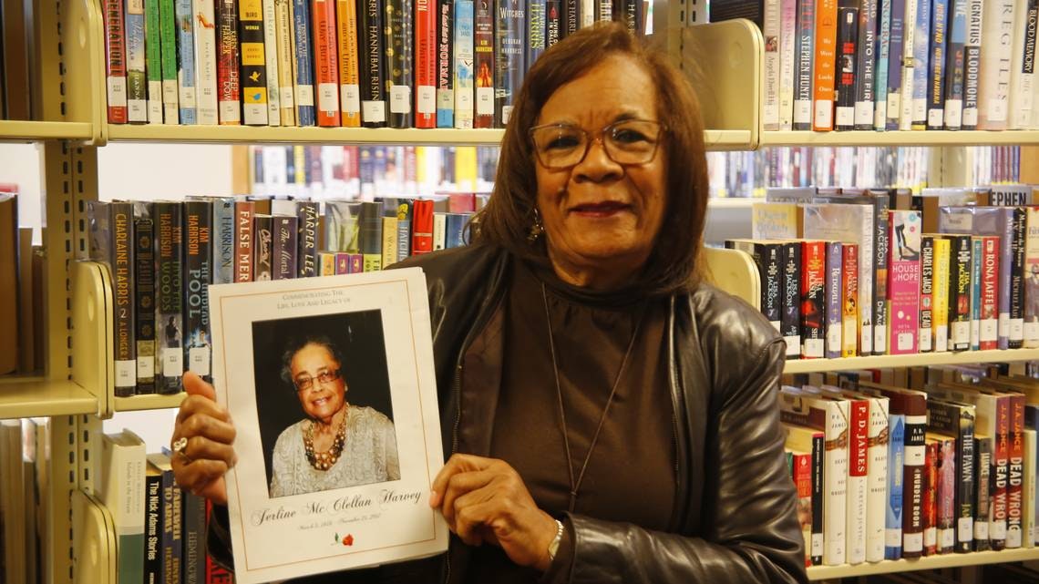 A woman holds a memorial program for Justine McClellan Harvey in a library, surrounded by bookshelves.