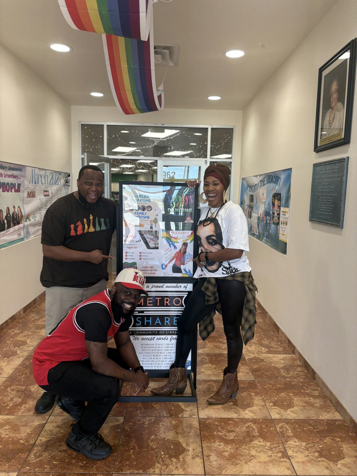 Three people posing happily in a hallway with a rainbow decoration and a poster about library events.