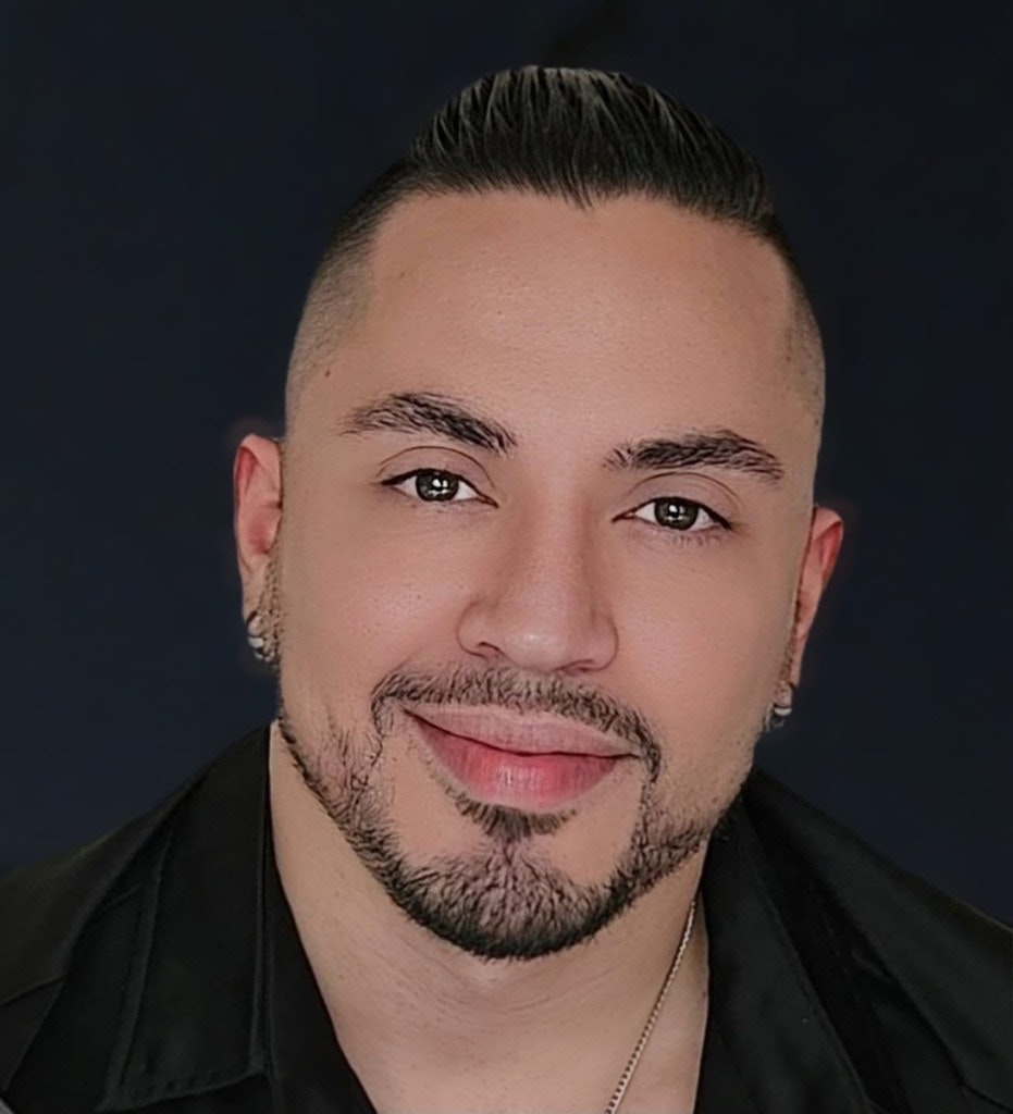 A person with short hair, earrings, and a beard, smiling, wearing a black shirt against a dark background.