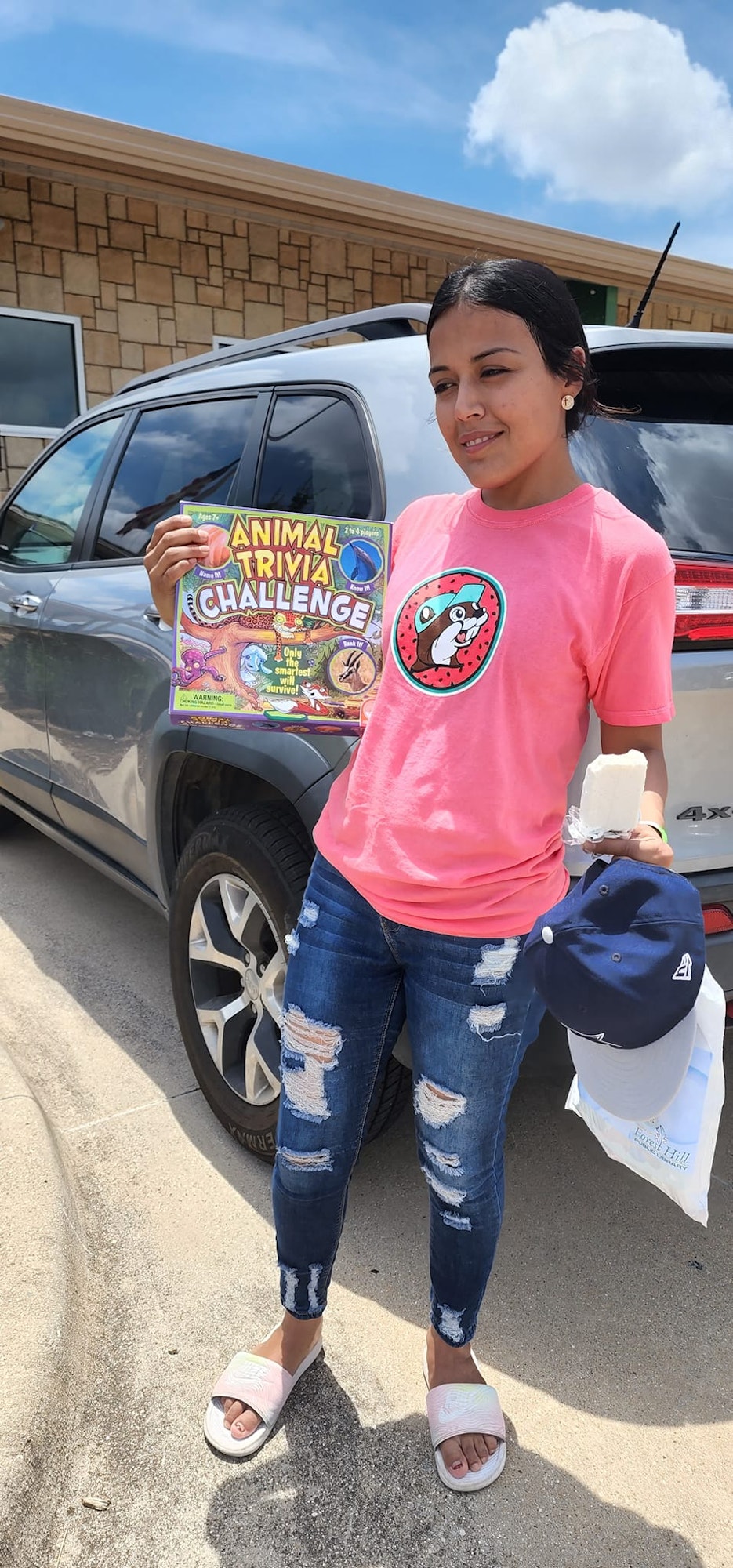 A person in a pink shirt holds an "Animal Trivia Challenge" box, a popsicle, and a cap, standing next to a car.