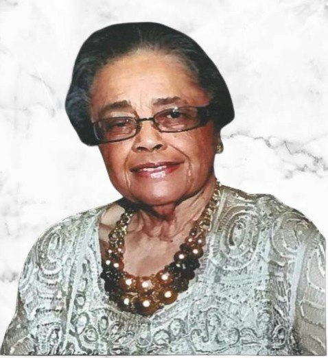 An older woman with glasses, wearing a patterned top and beaded necklace, against a marble-like background.