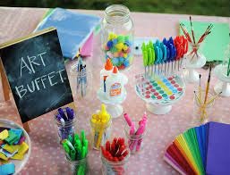Art supplies displayed on a table with a sign reading "Art Buffet"; includes markers, paper, glue, and colorful objects.
