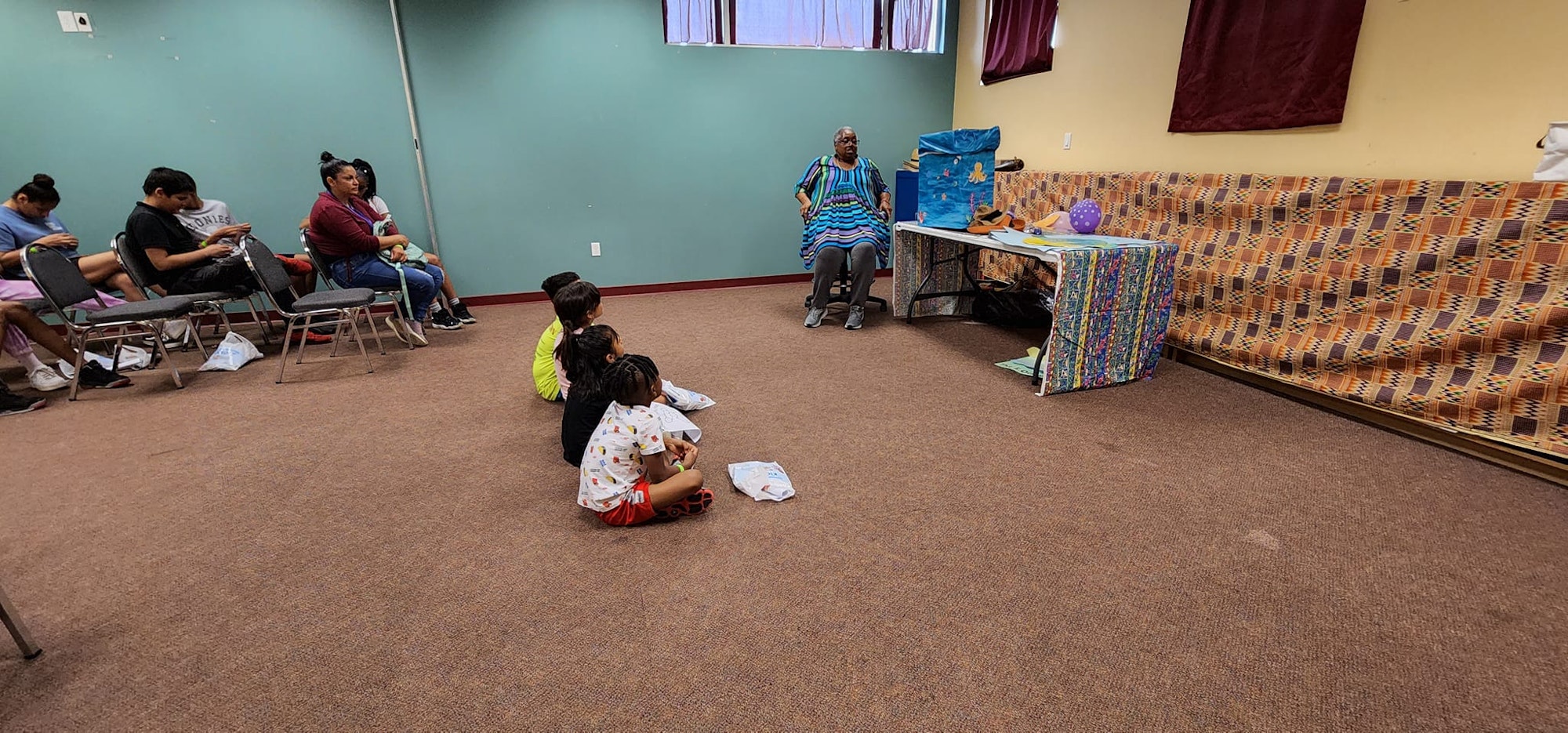A small group of children sit on the floor while adults sit on chairs in a room with decorated tables.