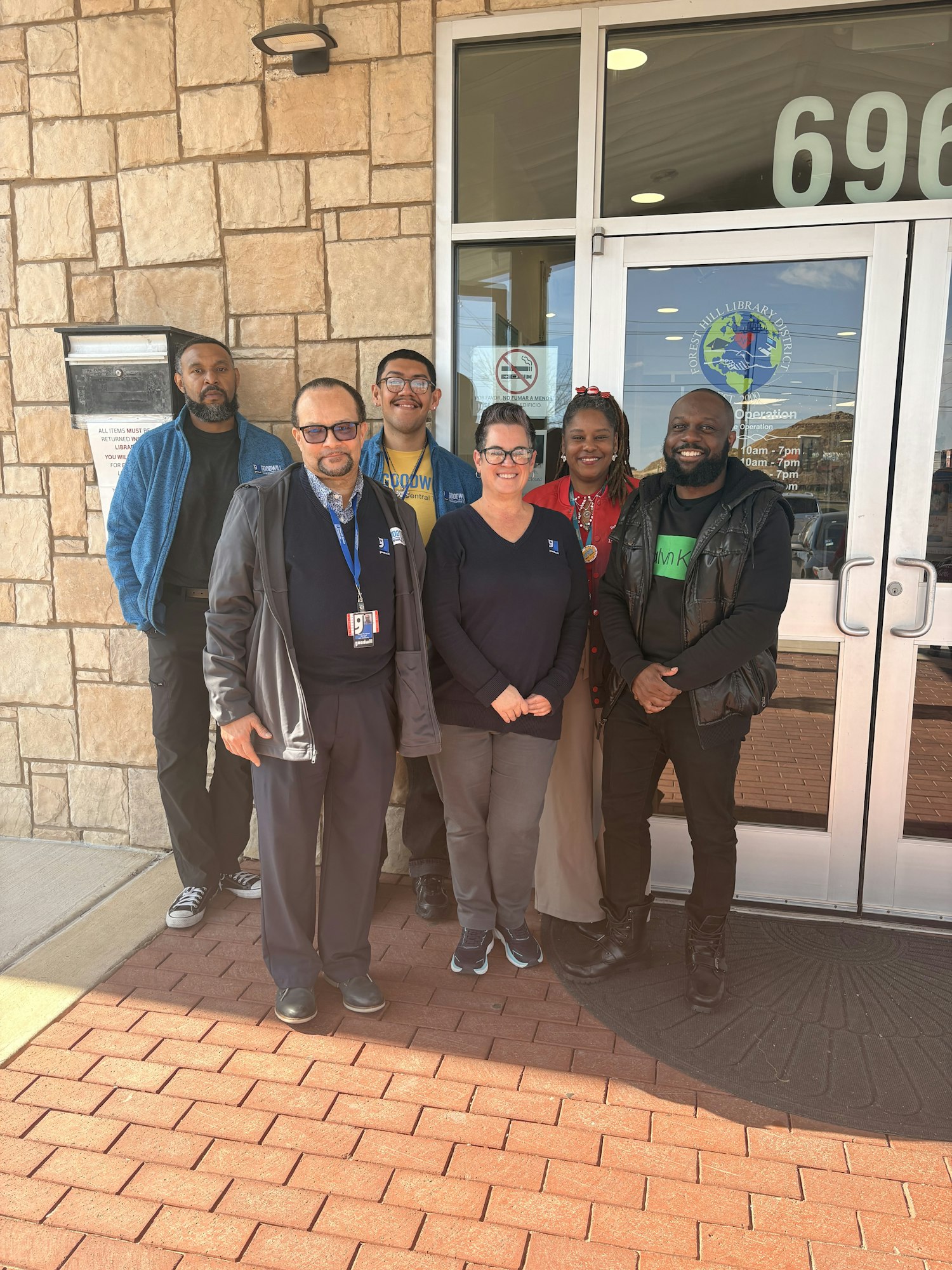 A group of six people posing in front of a building with a stone facade, likely a library or community center.