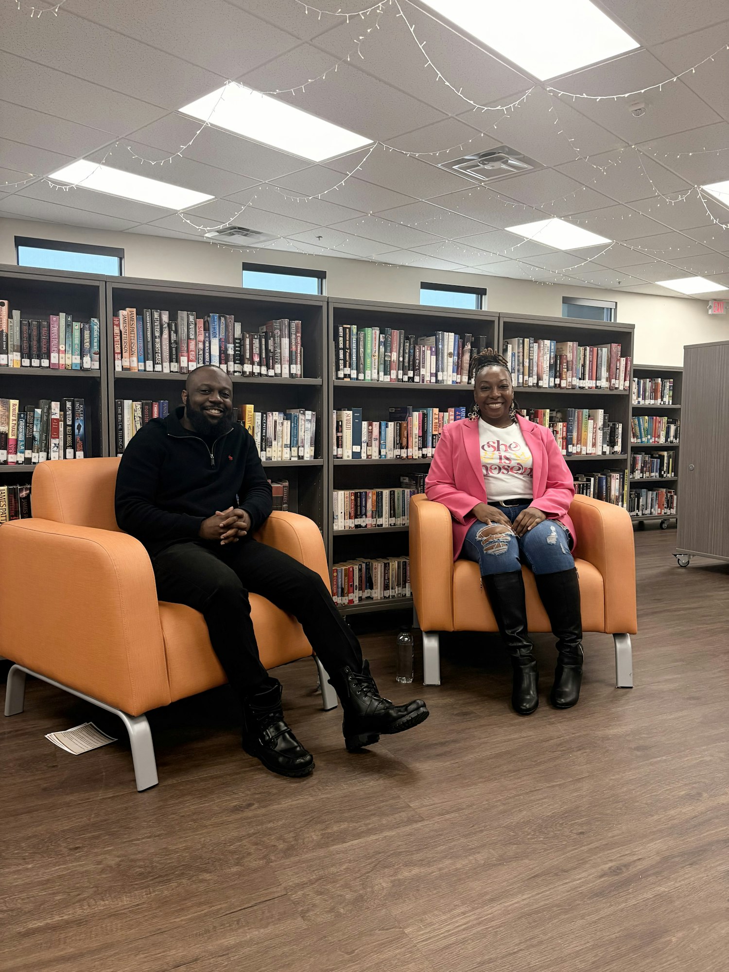 Two people are seated in orange chairs in a library, surrounded by bookshelves. They appear to be engaged in a discussion.