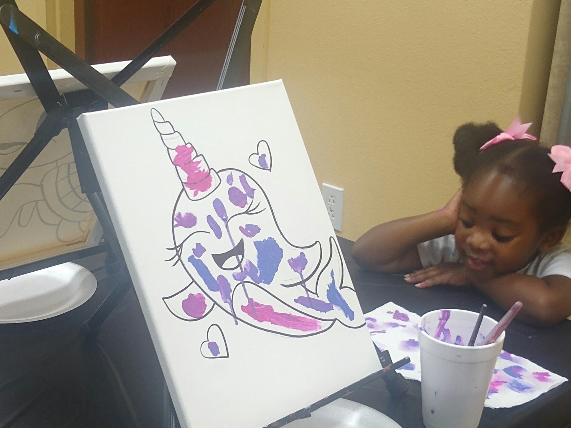 A child with pink bows is painting a colorful unicorn on a canvas with purple and pink paint.