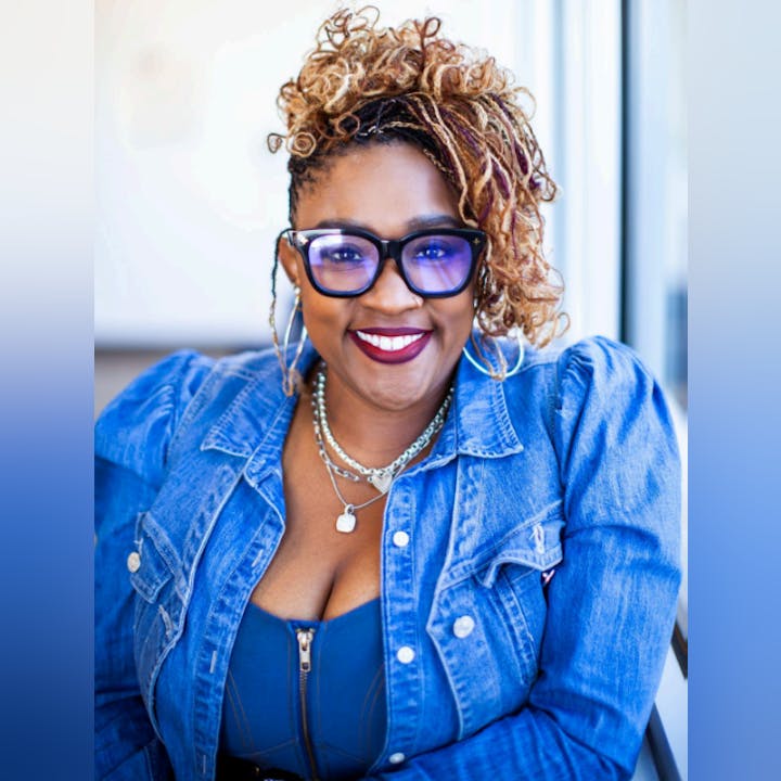 A smiling person wearing glasses and a denim jacket, with styled hair and jewelry.