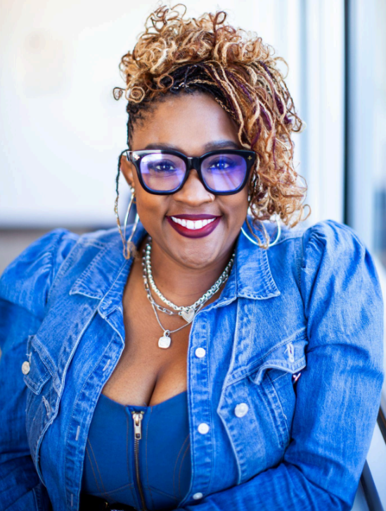 A smiling person wearing glasses and a denim jacket, with styled hair and jewelry.