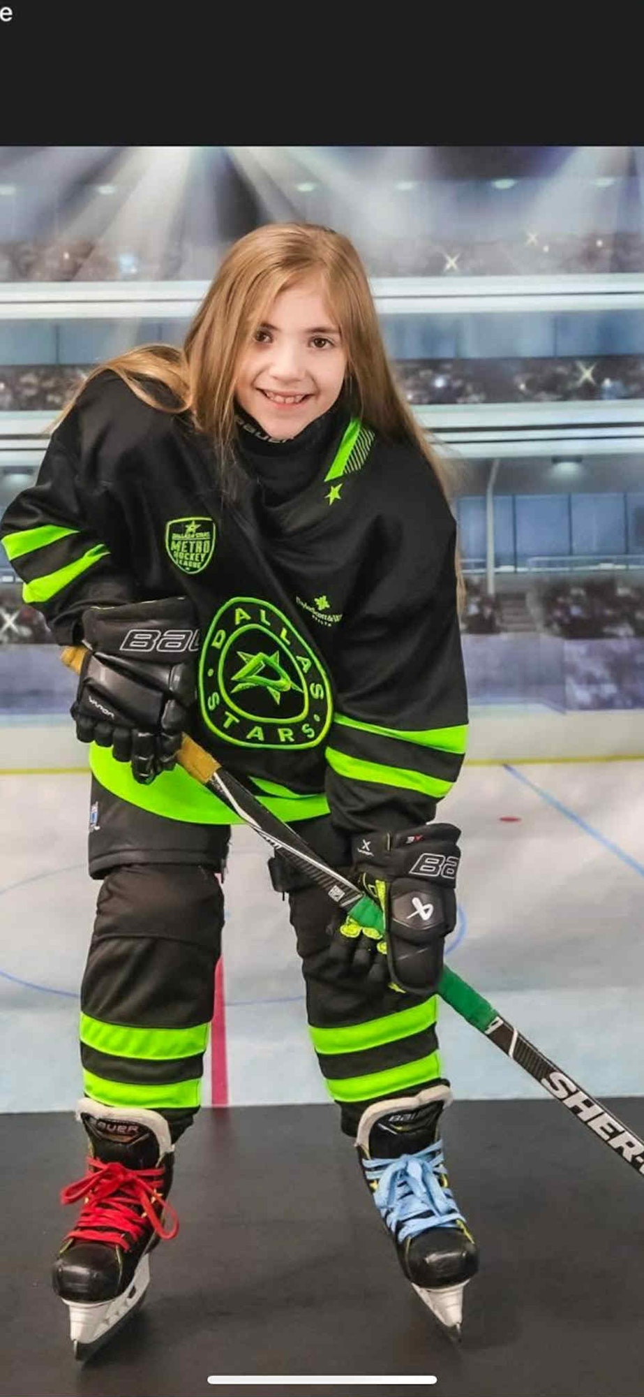 A child in a Dallas Stars hockey uniform holding a stick, wearing mismatched skate laces—one red, one blue.