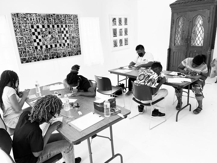 A group of people sitting at tables, drawing or writing, in a room with artwork on the walls.