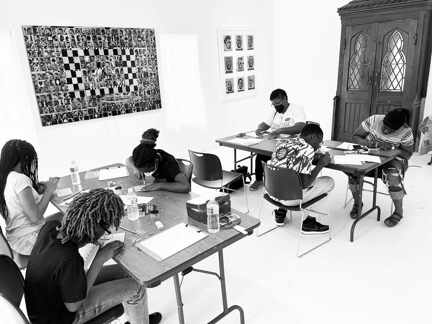 A group of people sitting at tables, drawing or writing, in a room with artwork on the walls.