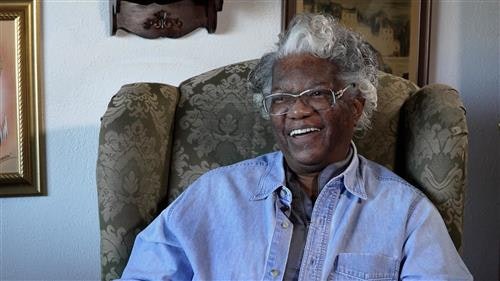 An older person with glasses is sitting in an armchair, smiling, wearing a light blue shirt.