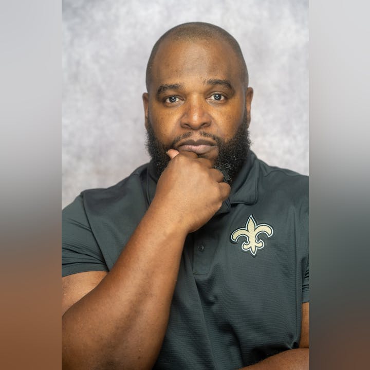 A man with a beard, wearing a black shirt with a fleur-de-lis emblem, rests his chin on his hand.