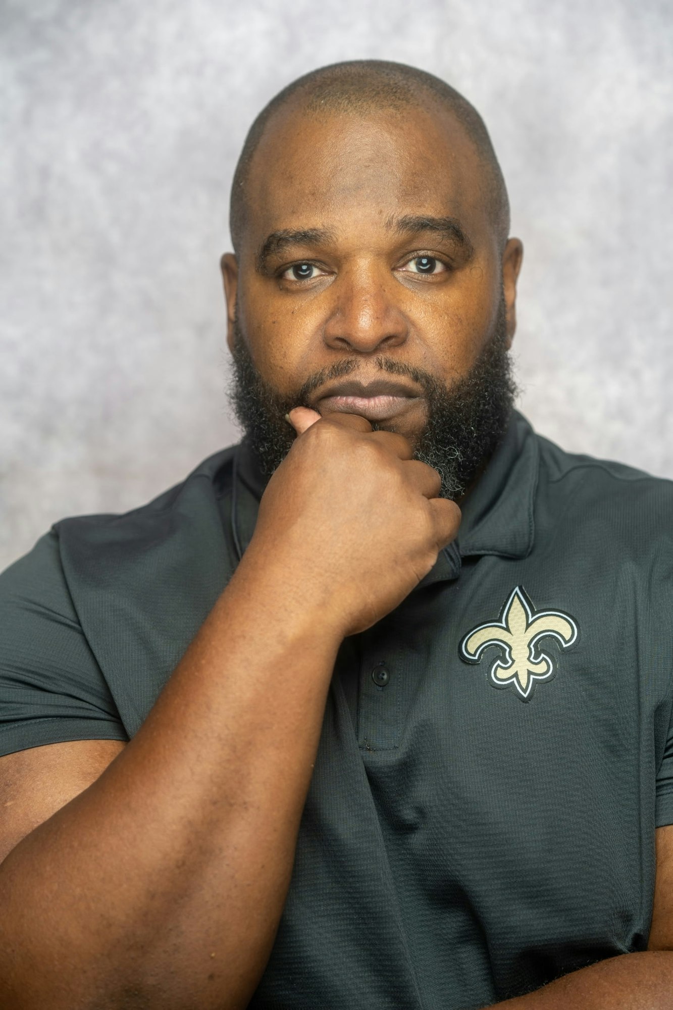 A man with a beard, wearing a black shirt with a fleur-de-lis emblem, rests his chin on his hand.