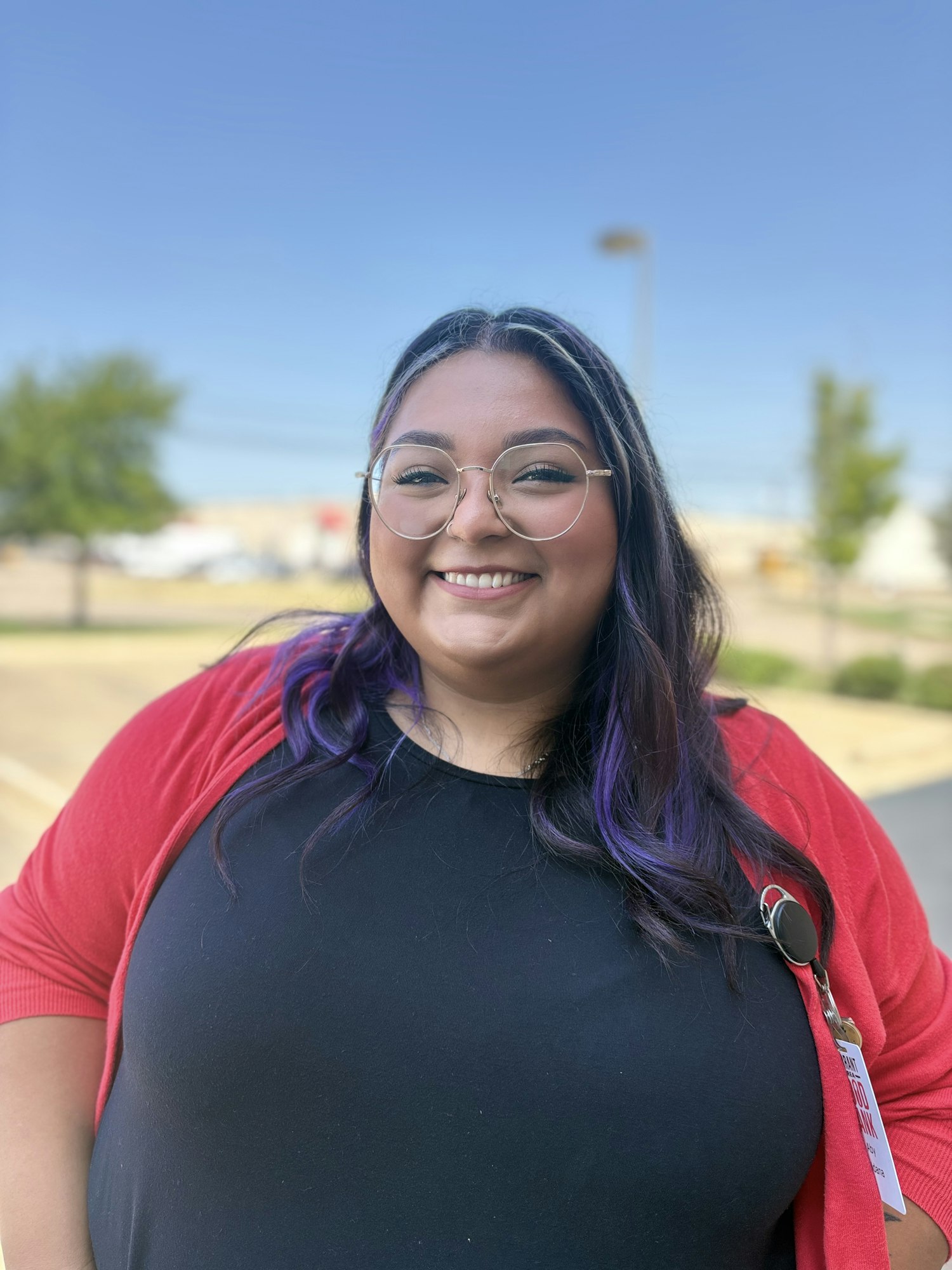 A person wearing glasses and a red cardigan, smiling outdoors with a clear blue sky in the background.