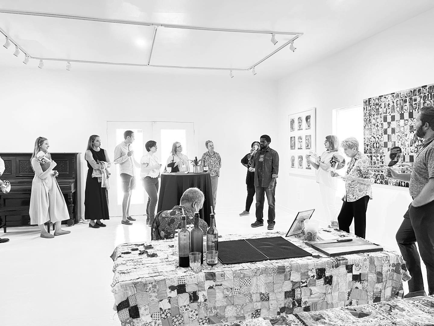 People in a gallery space looking at art, with a table covered in quilts and bottles in the foreground.