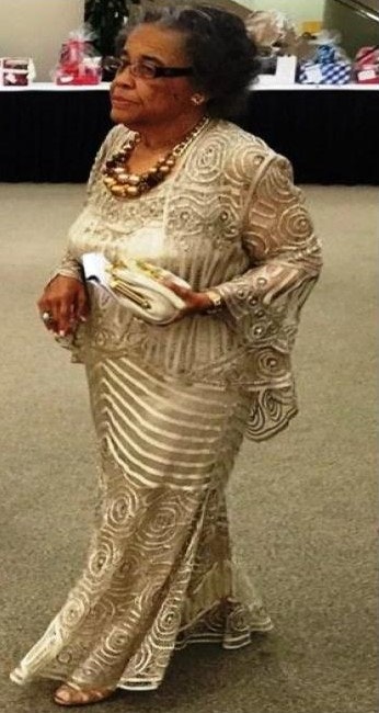 An elegant woman in a decorated gown holding a clutch, wearing a necklace, walking indoors.