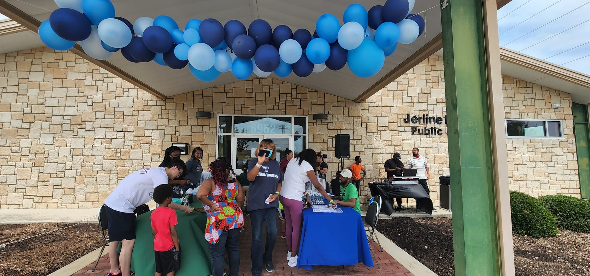 People gather at an event under blue and white balloons, with a table setup at a public building entrance.