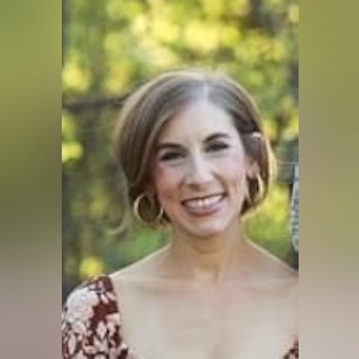 A person smiling outdoors with trees in the blurred background, wearing a floral outfit and hoop earrings.