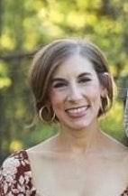 A person smiling outdoors with trees in the blurred background, wearing a floral outfit and hoop earrings.
