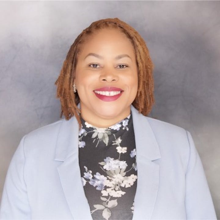 The image shows a smiling woman with dreadlocks, wearing a floral top and light blazer, against a gray background.