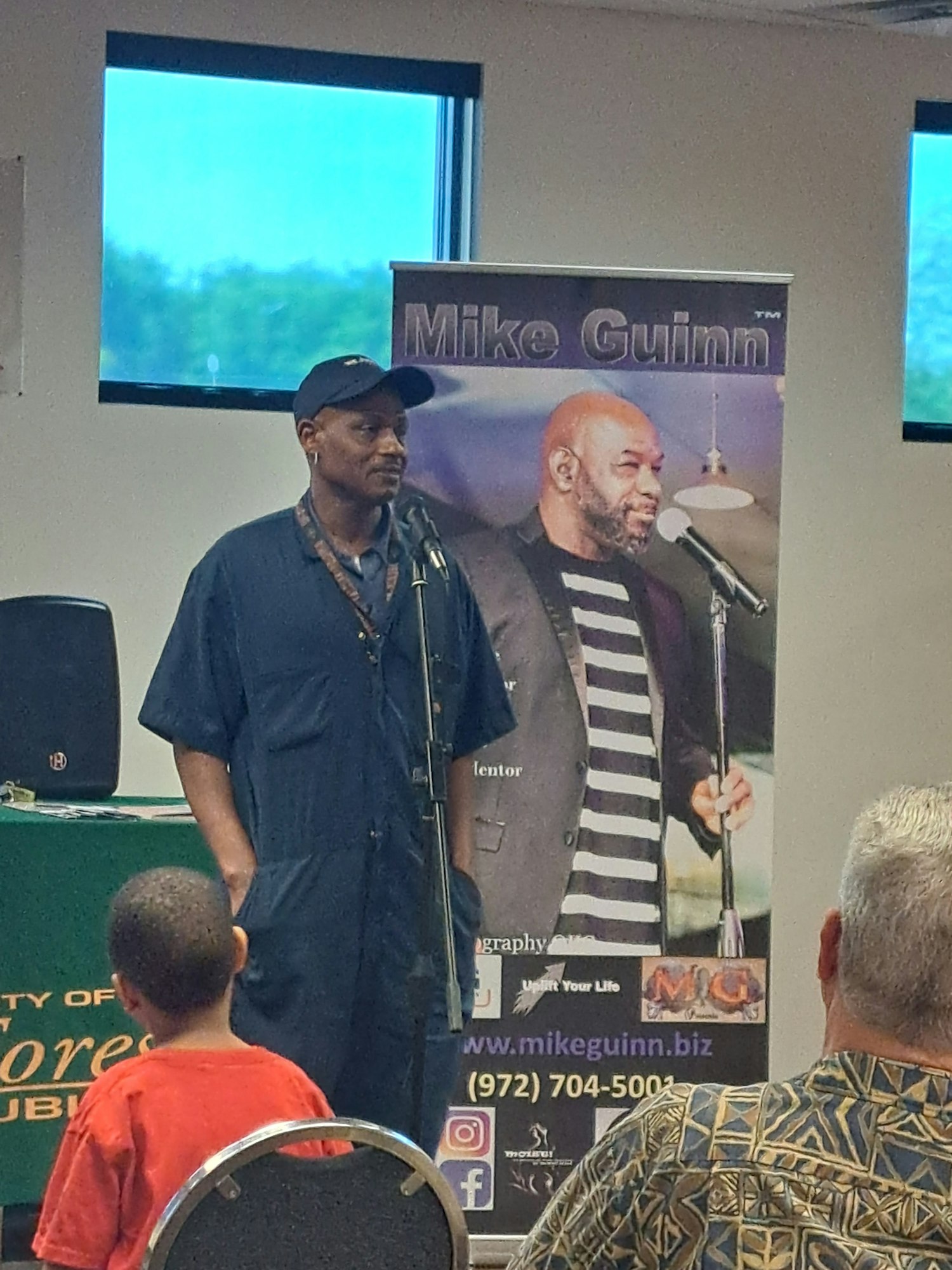 A man speaks into a microphone. A banner with "Mike Guinn" and a photo of another man is behind him. Audience members are seated.
