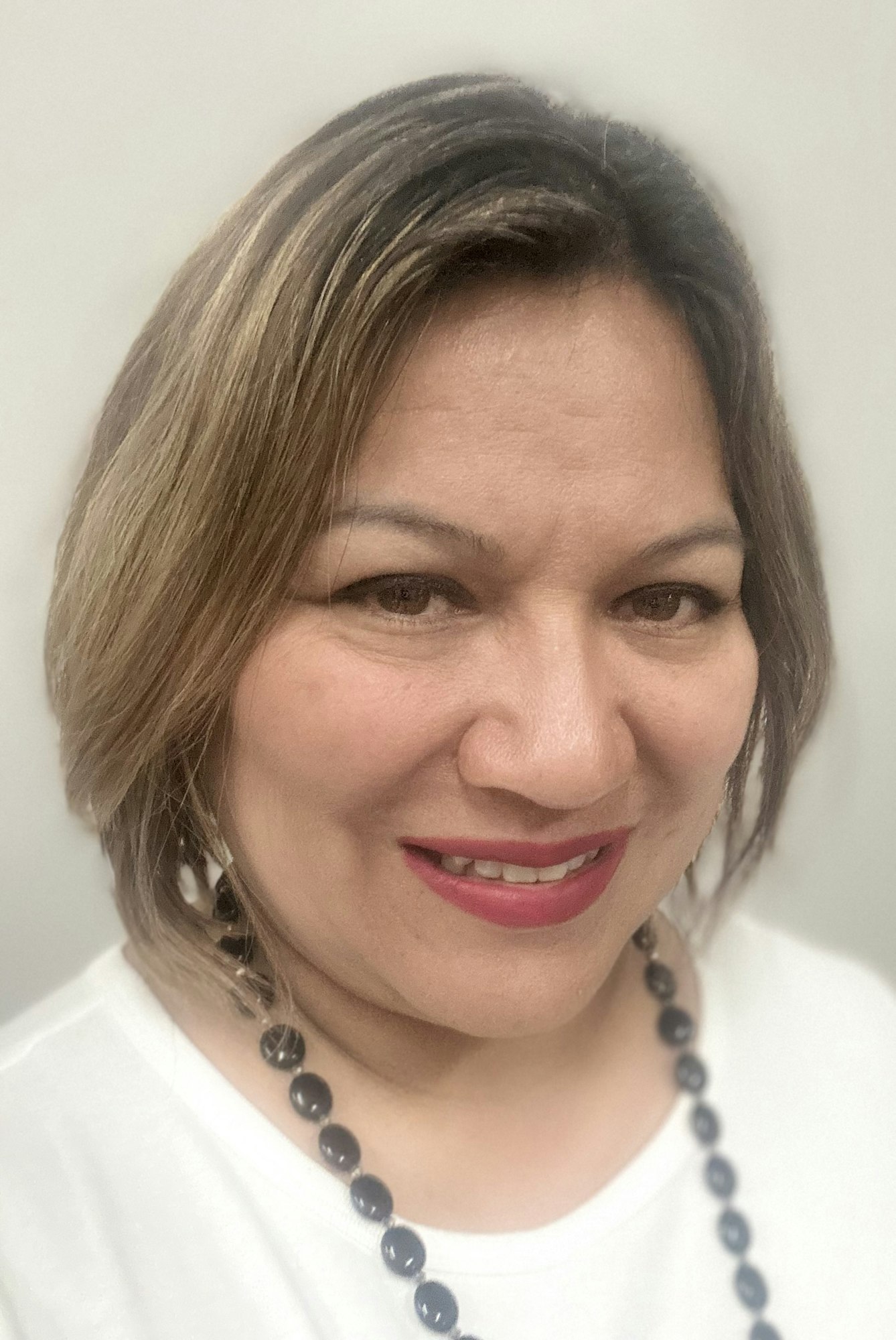 A close-up of a smiling woman with medium-length hair, wearing a white top and a dark necklace, set against a light background.