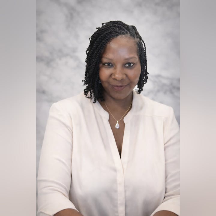 A woman with braided hair smiles, wearing a light blouse and a necklace, set against a neutral background.