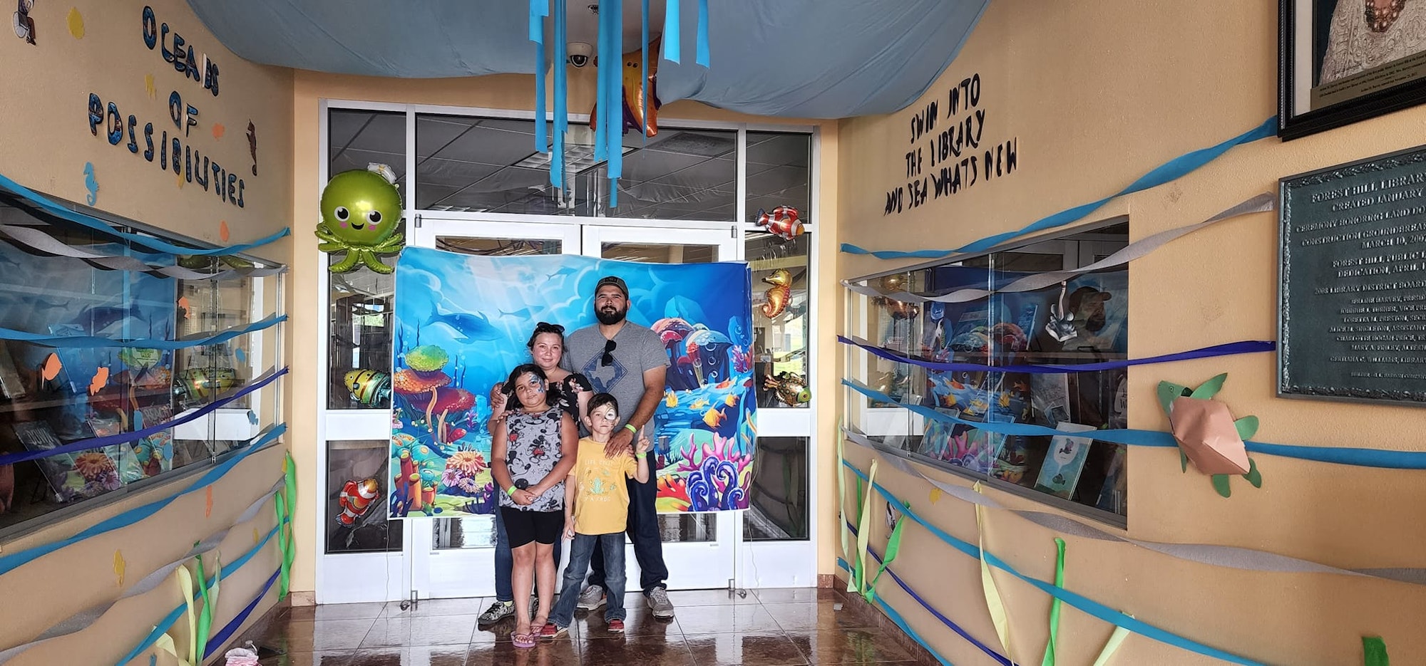 A family poses in front of an underwater-themed library entrance with "Ocean of Possibilities" decor, colorful streamers, and sea creatures.
