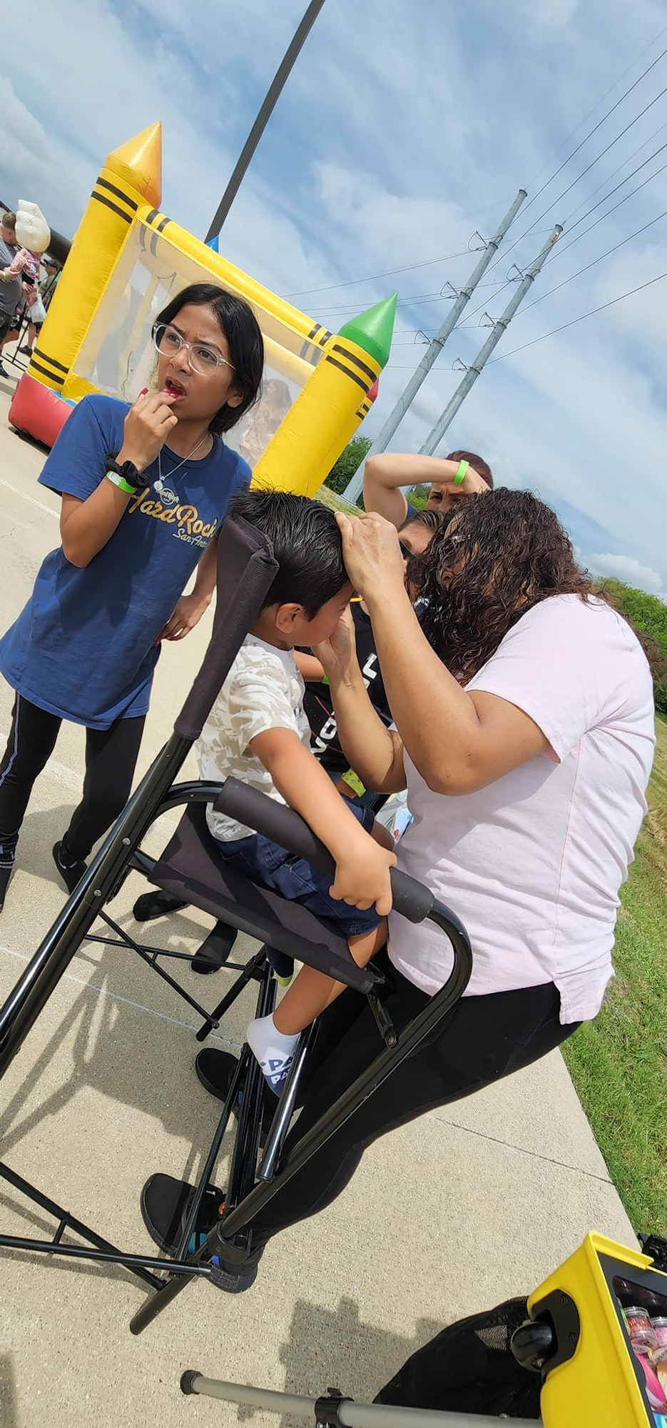A woman styles a child's hair outdoors, while another child watches. A yellow inflatable is in the background.