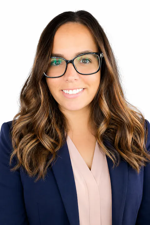 The image shows a woman with wavy hair wearing glasses and a professional outfit, smiling against a white background.