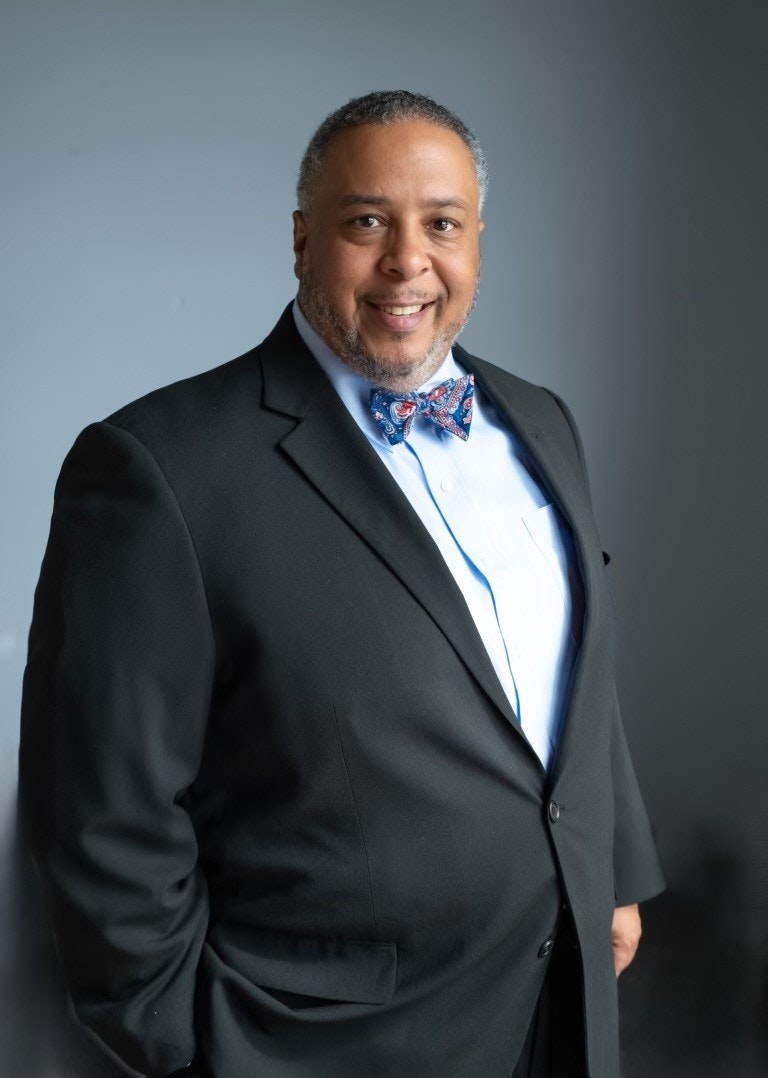 A person smiling, wearing a black suit with a blue bow tie, against a plain background.