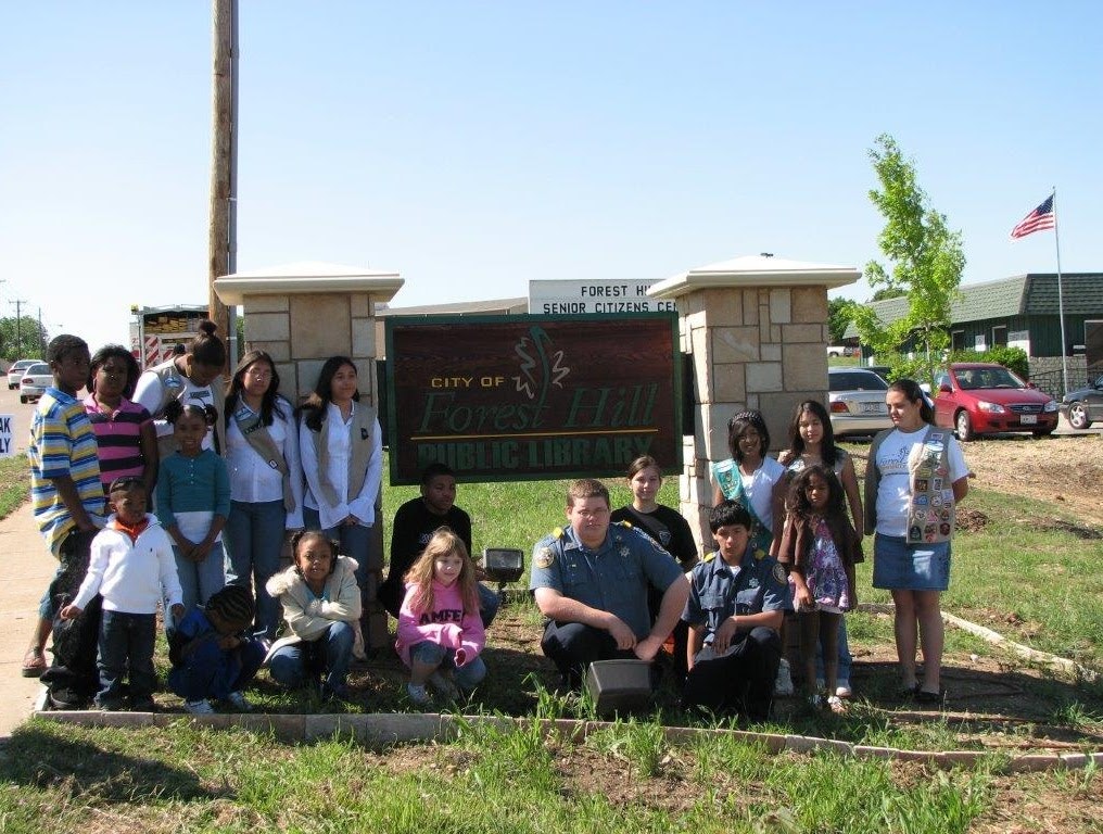 A group of people, including children and uniformed individuals, stand in front of a "Forest Hill Public Library" sign.