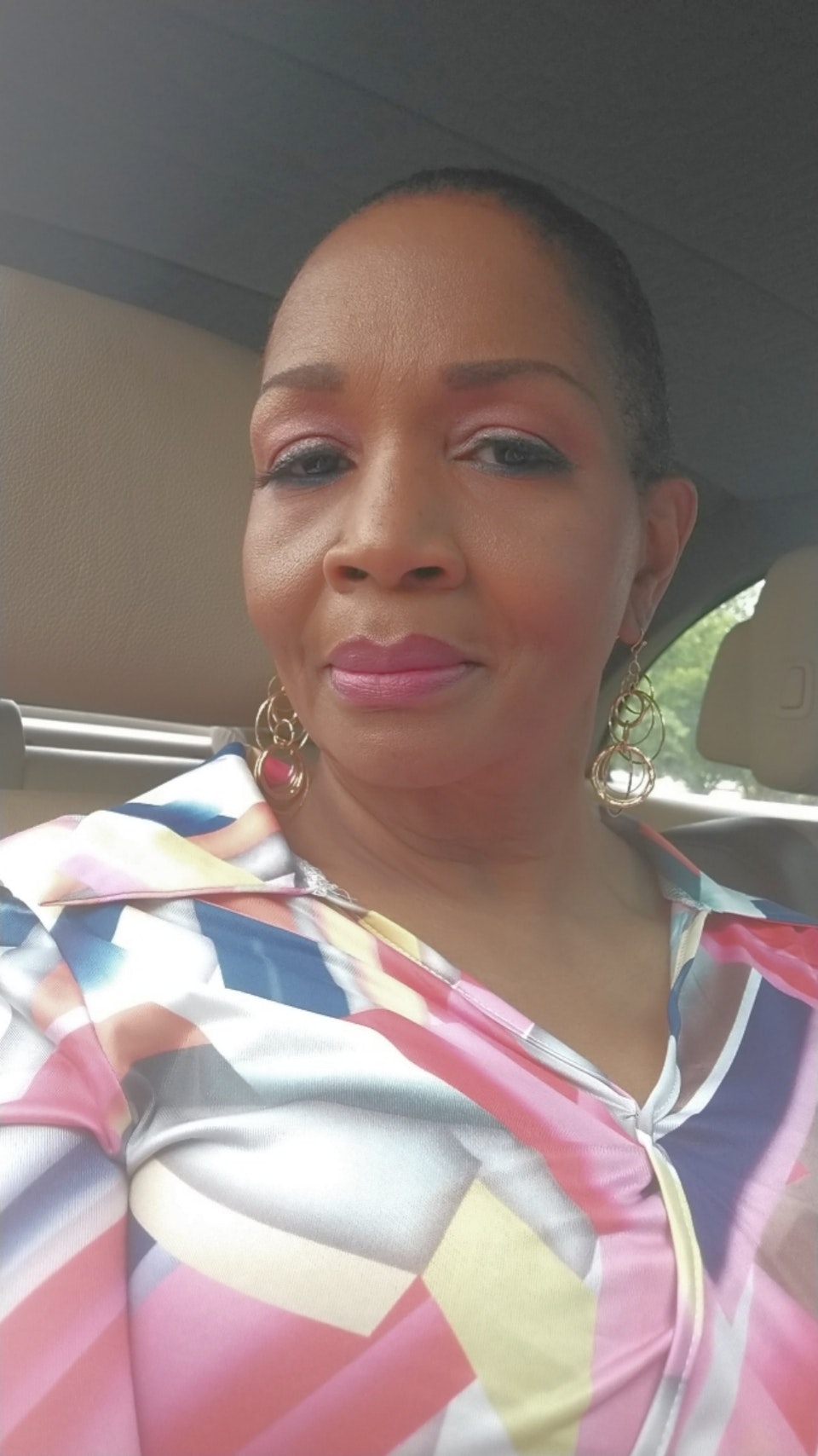 A person sitting in a car wearing a colorful geometric-patterned shirt and gold hoop earrings.