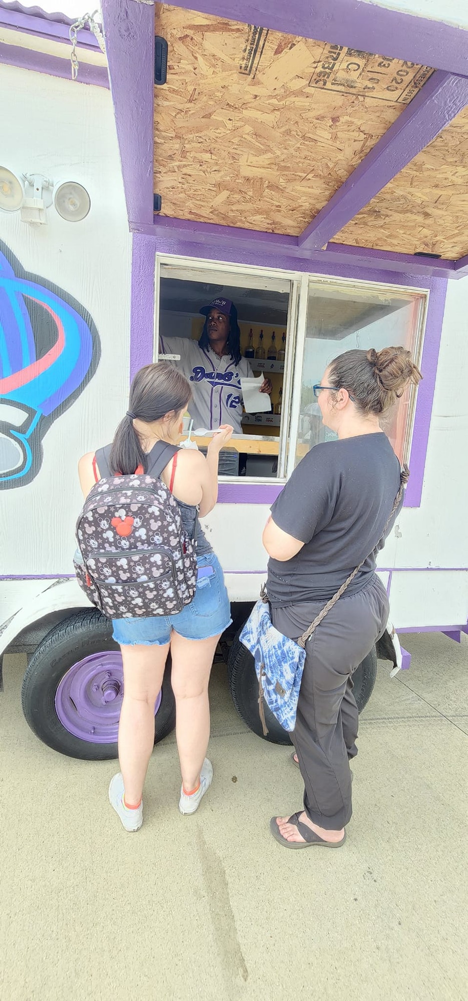 Two people order from a vendor at a food truck with a purple and white design.