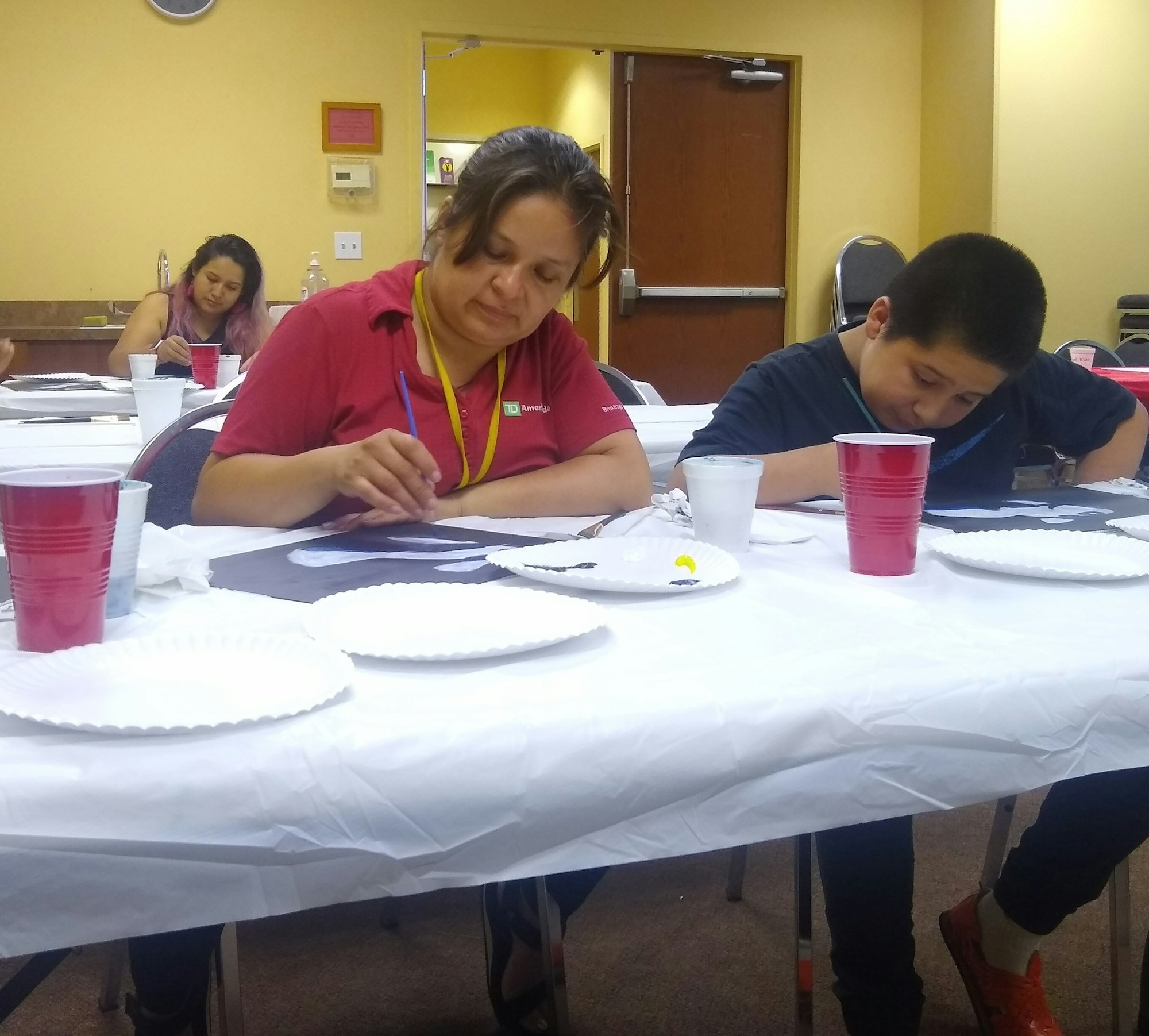 People painting at tables with art supplies and drinks in a room.