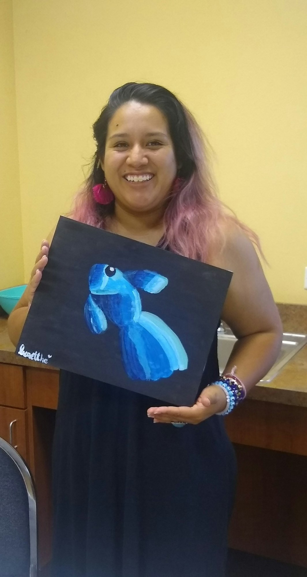 A person holding a painting of a blue fish, smiling, in a room with yellow walls.