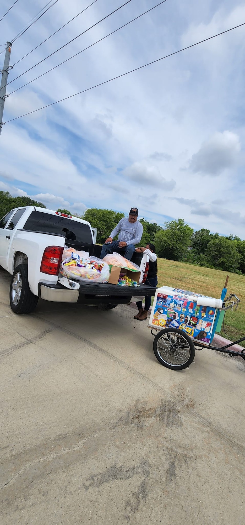 A person sitting on a truck bed with bags and boxes of snacks; a colorful ice cream cart attached to the truck.