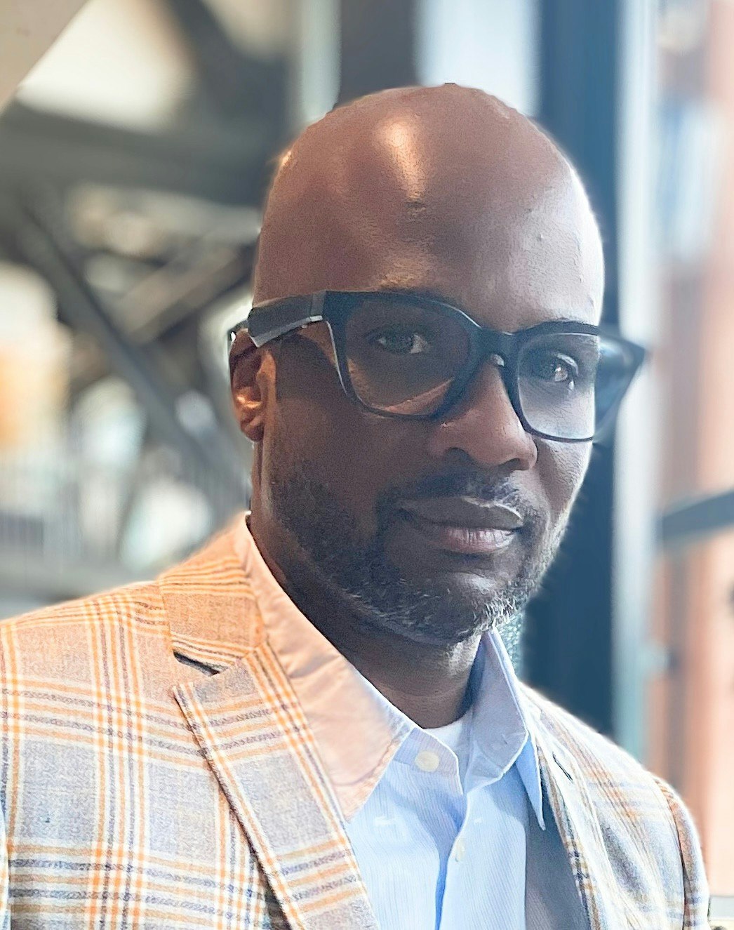 A person with glasses, wearing a plaid jacket and shirt, is looking at the camera.