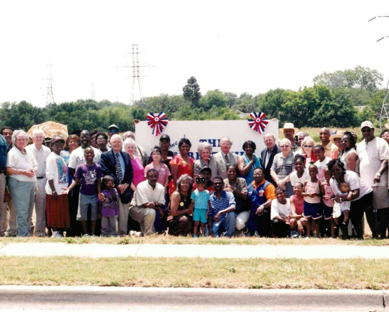 A large group of people posing together outdoors, with some holding a sign. Trees and power lines are in the background.