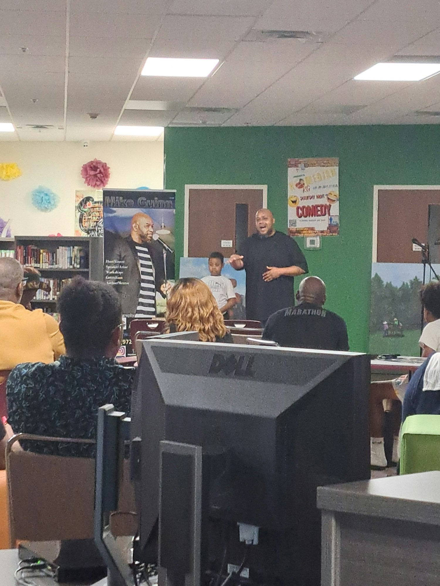 A man speaks to an audience in a room with posters and computers, next to a child and a large banner showing a microphone.